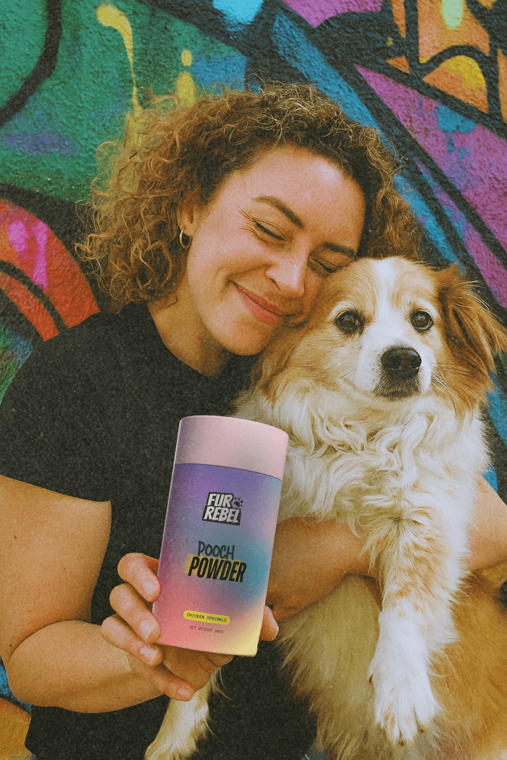 Woman holding a dog and a container of Fur Rebel pooch powder dog supplement powder against a colorful graffiti wall.